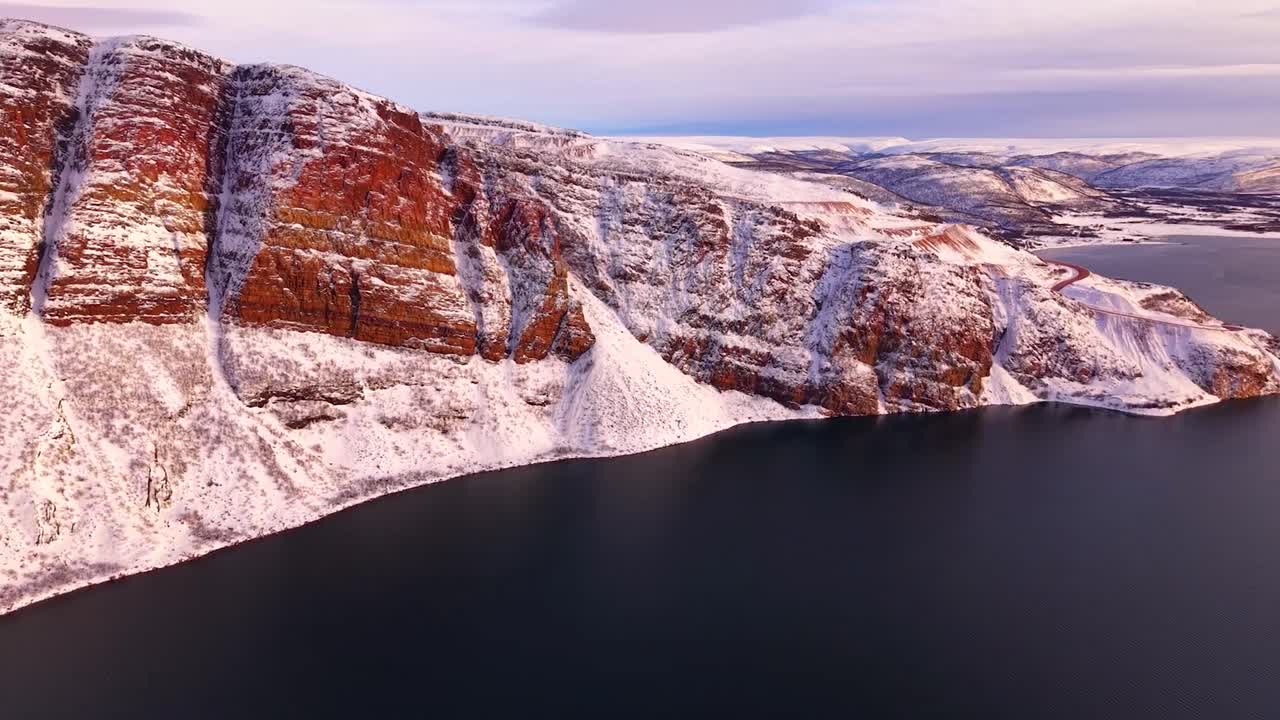 Aerial drone footage view of steep brown colored and white snow covered Giemaš mountain in nordic Norway Arctic during a sunny day while sun sets or rises with orange golden colors. Barents sea also.