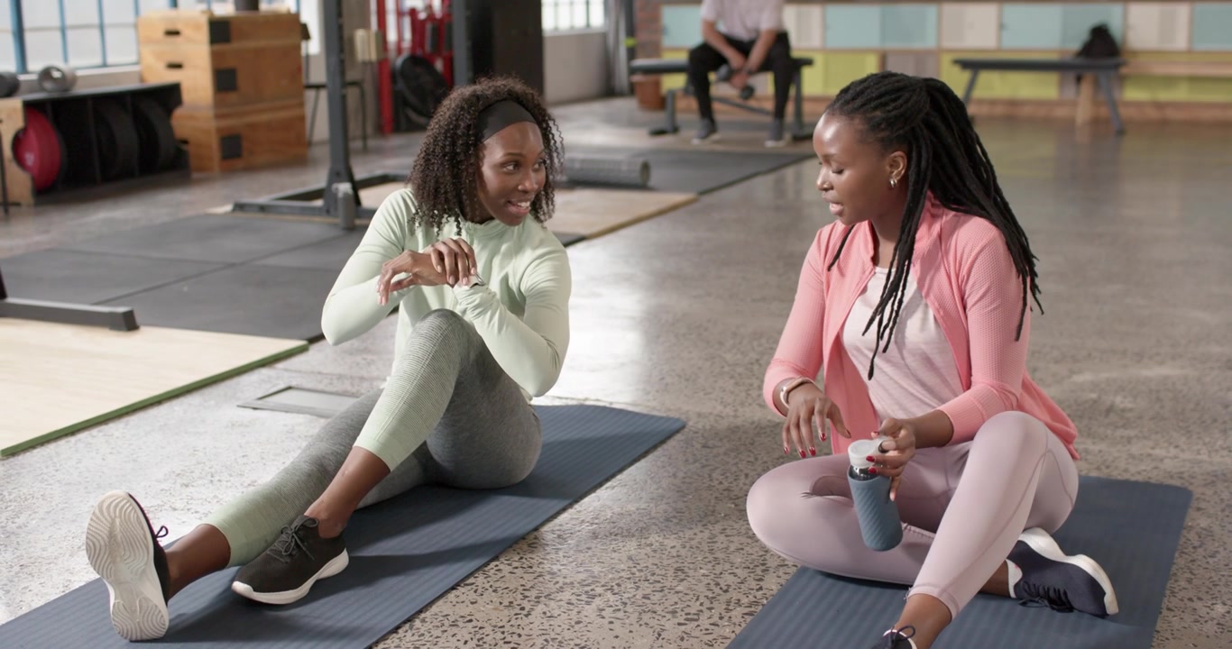 African American women sitting on mats in gym in activewear glancing at watches and comparing stats