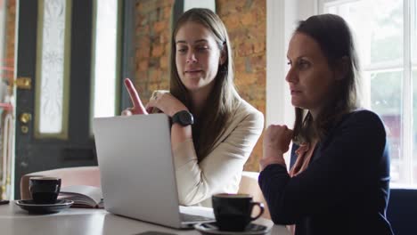 dos mujeres de negocios caucásicas sonrientes sentadas en una mesa con café, usando una computadora portátil y hablando,
