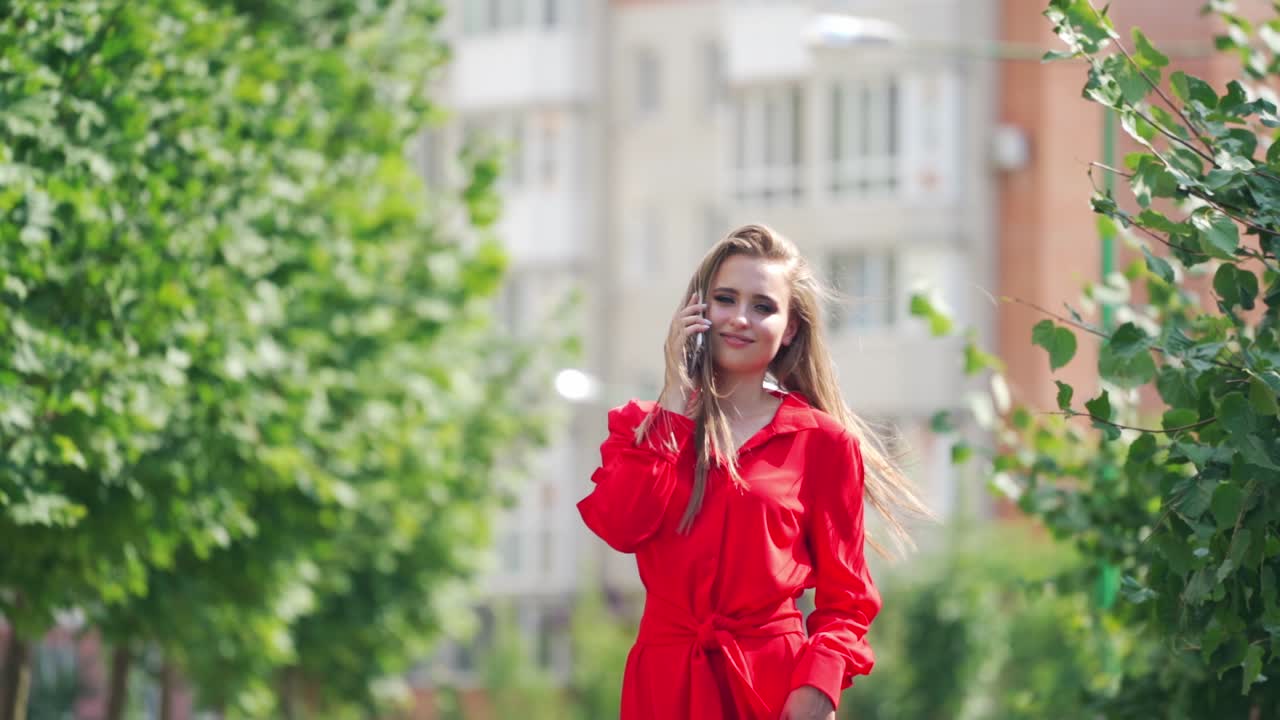 Incredible woman in the city. Portrait of a beautiful young female who is talking on a mobile phone. Gorgeous smiling lady in red dress outdoors in summer.