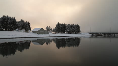 Reflection-of-trees-and-building-in-snowy-landscape-on-calm-fjord-water-surface-at-sunset