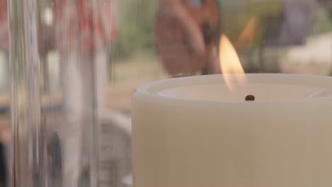 primer plano de una vela encendida en una linterna, alguien tocando la guitarra en el fondo