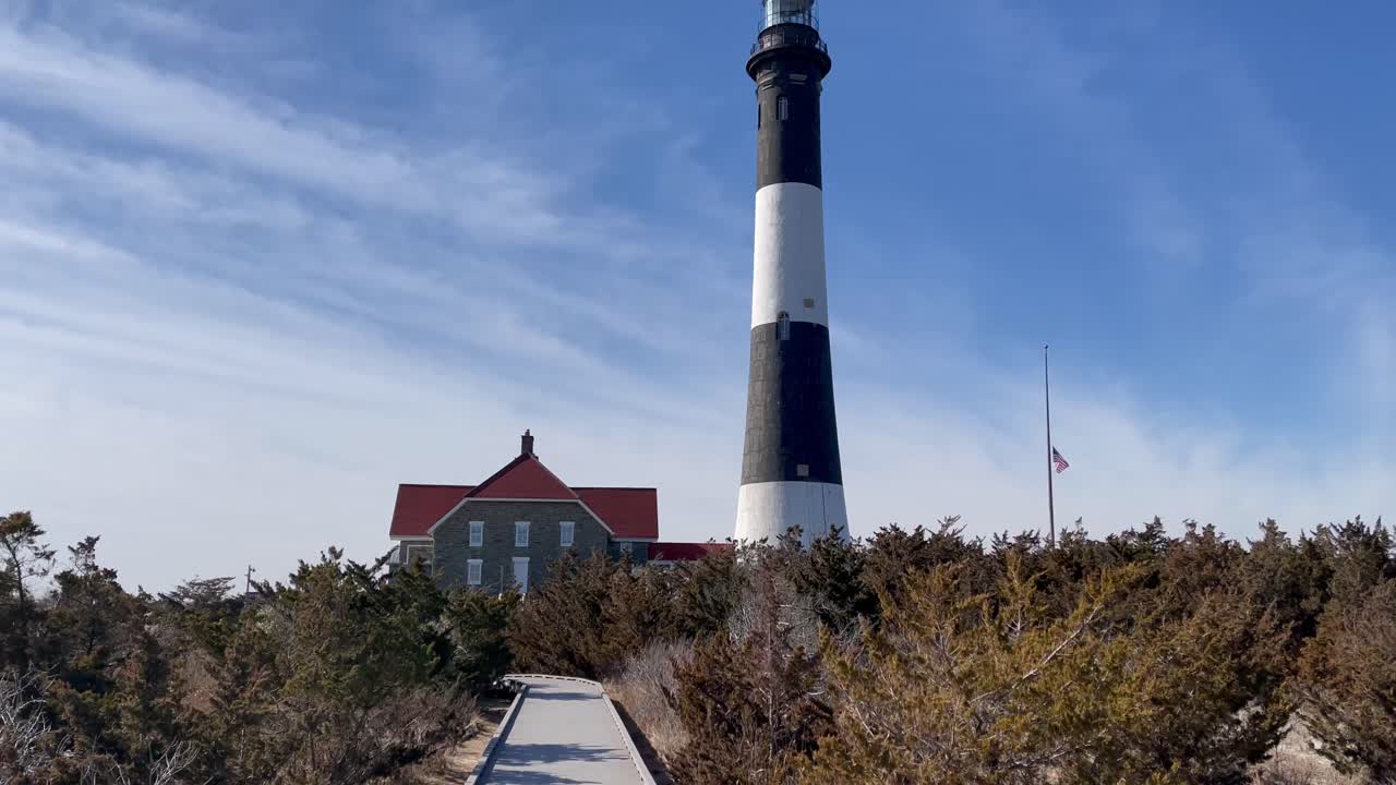 Fire Island Lighthouse tall black and white tower stands behind red-roofed station, wooden boardwalk path leads through dune forest, bright sky and historic coastal landmark in New York