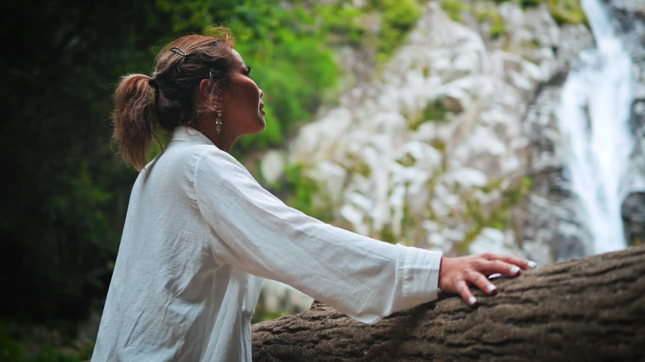 mujer de camisa blanca con el cabello atado hacia atrás se apoya en una barandilla de madera, mirando hacia arriba a una cascada en un bosque verde y exuberante enfatizando la belleza natural de la cascada y el entorno sereno