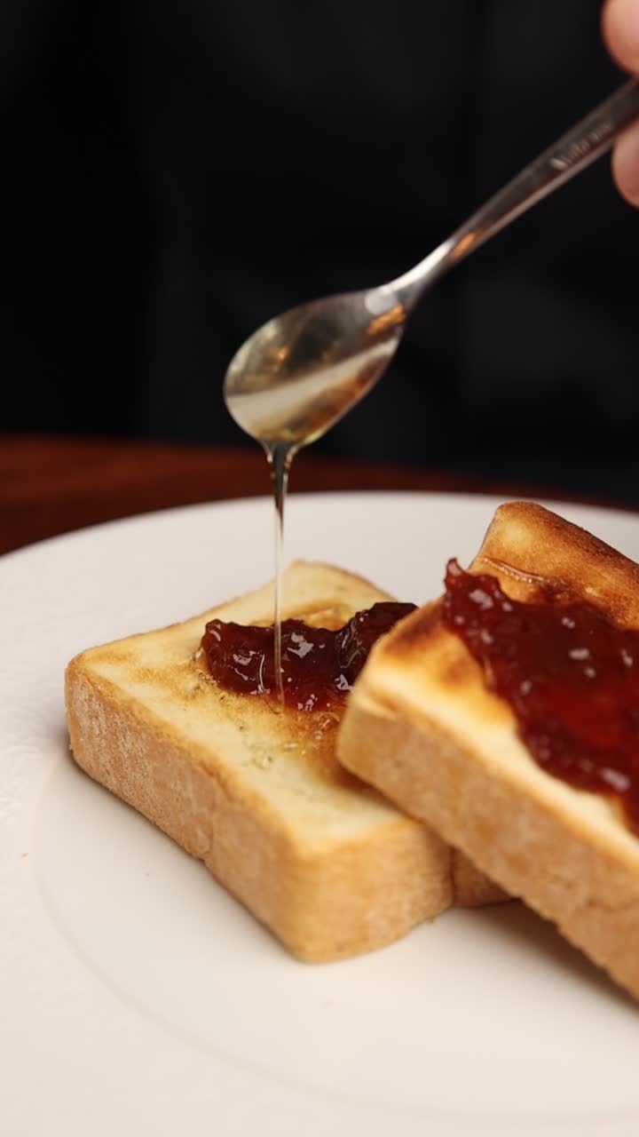 Drizzling Golden Honey onto Toast with Red Jam on a White Plate