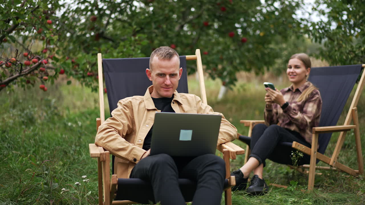 Young couple working remotely out of office. Man uses his laptop and woman speaks on the phone. Garden backdrop.