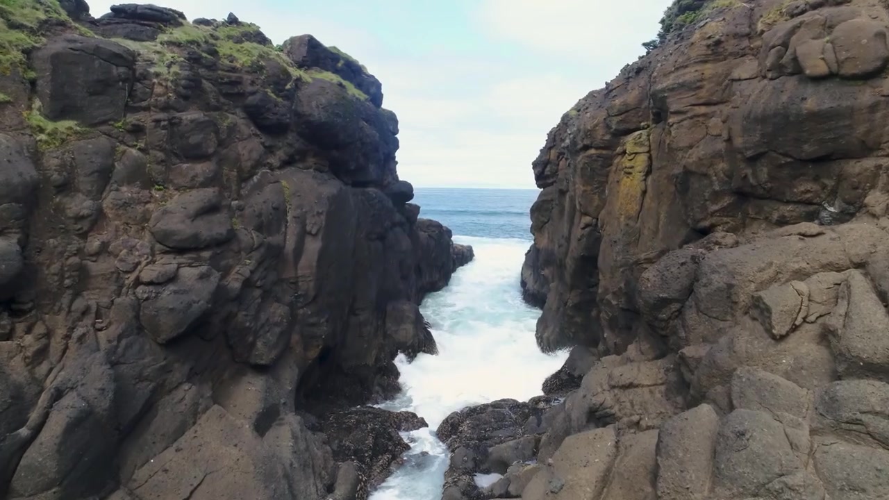 Low-flying wide-angle drone shot skimming over water through a narrow rock channel at Whale Cove south of Depoe Bay on the Oregon Coast, where waves surge through the sea-cut opening.