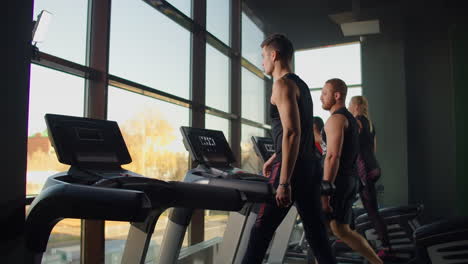 men walk on treadmills in the fitness room. a group of people walking on treadmills near a large panoramic window. start of fitness classes. young beautiful women and men in sportswear in cardio fitness room