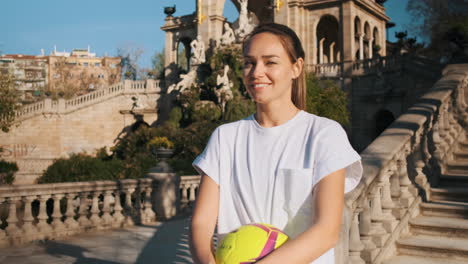 sporty woman with ball in hands happily looking at camera in city park.