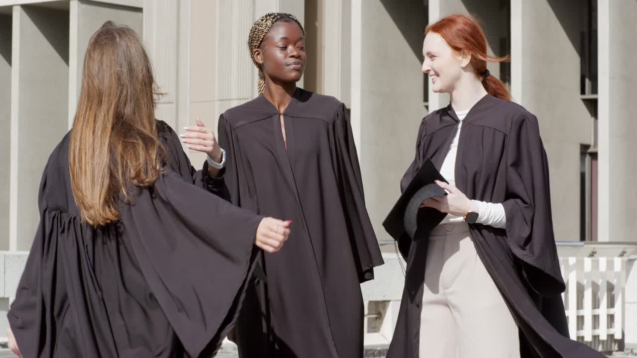 Diversas mujeres graduadas con toga y birrete animando, pelirroja levantando el birrete, caminando por la plaza