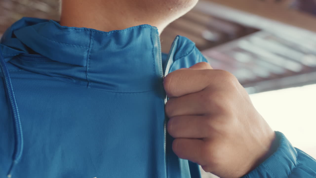 Primo piano di un atleta che chiude la cerniera della giacca blu prima della partita, con le mani che stringono la cerniera sotto un riparo con pioggerellina sul tessuto, cappuccio sollevato, profilo concentrato che rivela determinazione durante la preparazione per l'allenamento di calcio sul bagnato