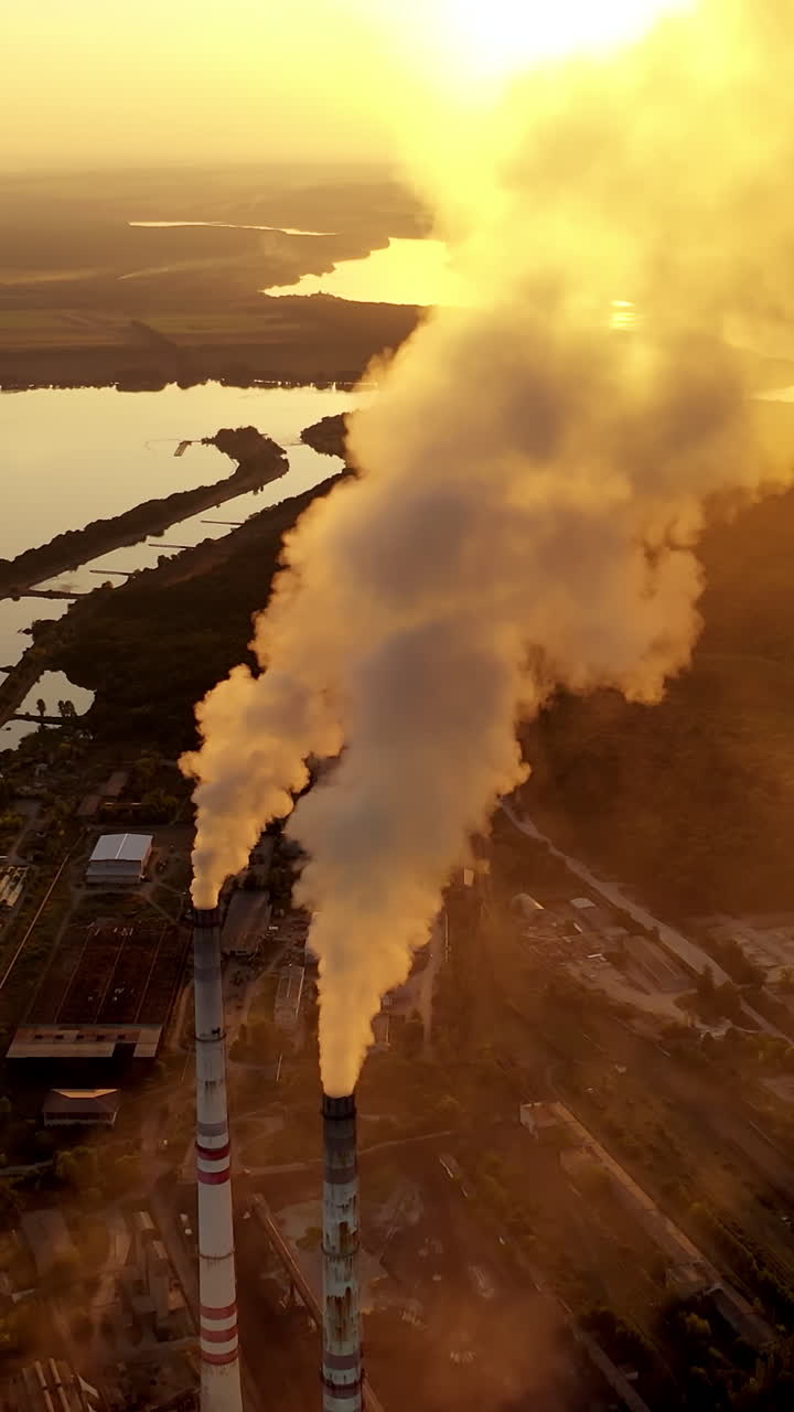 Smoking factory pipes at sunset. White smoke pours out of factory chimney in the air on the river background. Pollution, global warming concept. Aerial view. Vertical video
