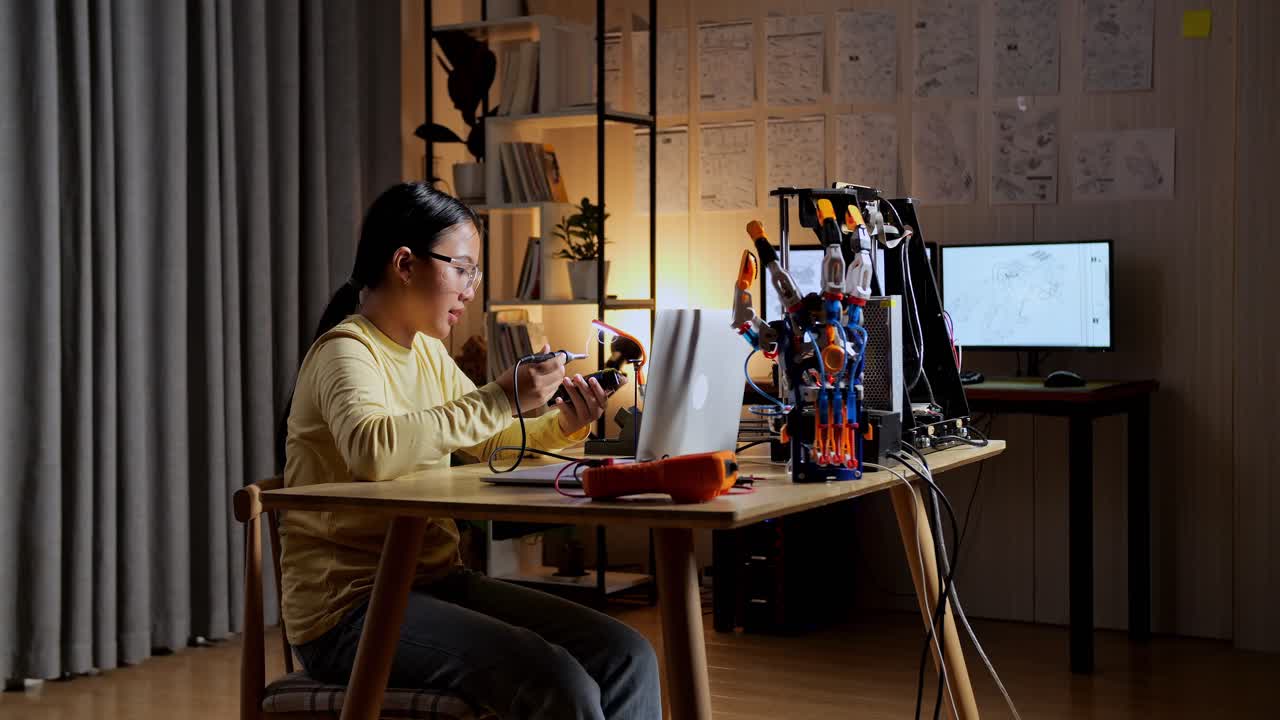 십대 아시아 소녀가 집에서 사이보그 손을 수리하는 동안 회로판을 들고 고치고 있습니다.