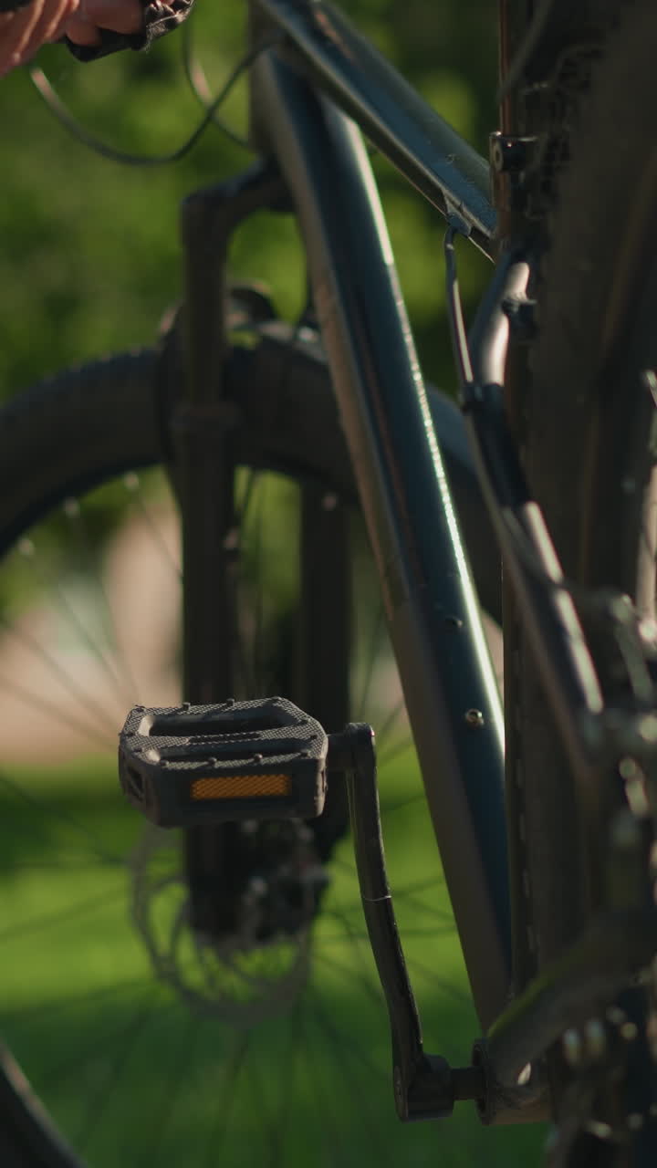 primer plano de una pierna de dama y una mano con guantes mientras elimina la bomba de aire verde del área del pedal de la bicicleta, con un fondo verde exuberante de árboles y un edificio lejano borroso