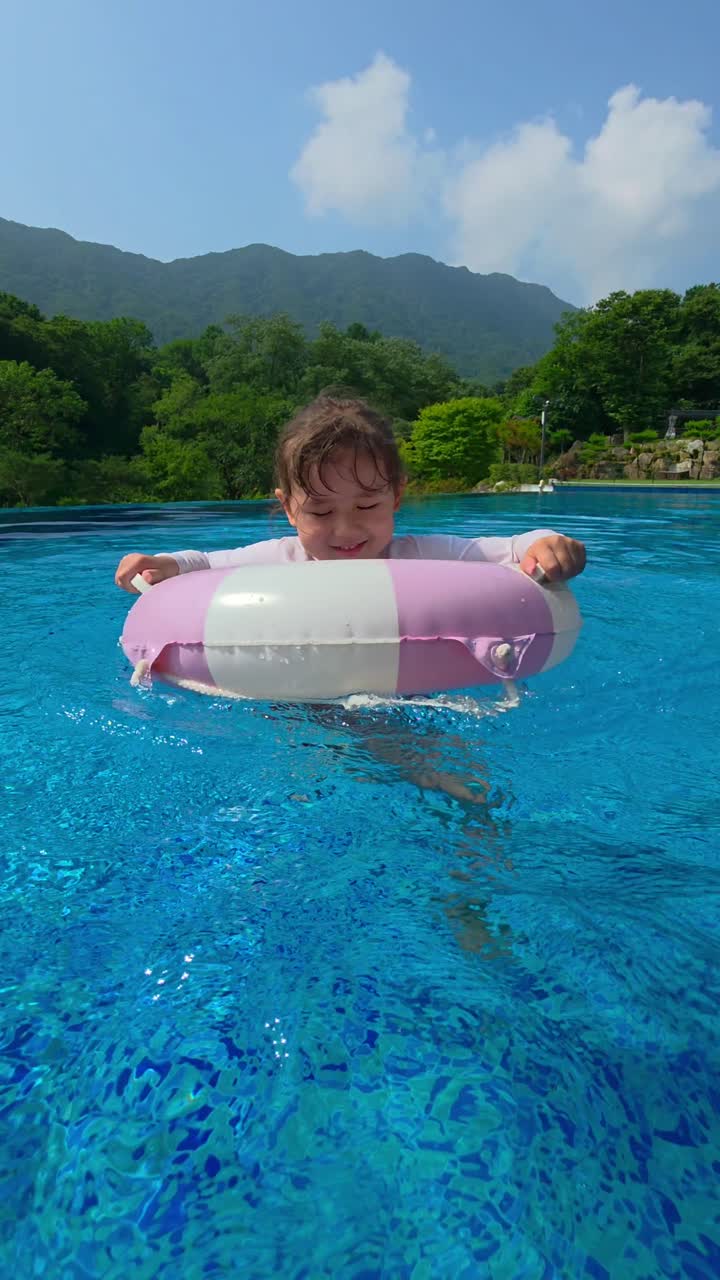 An excited 5-year-old mixed Korean girl spins around in her inflatable ring, splashing in a luxury infinity pool with a scenic green mountain landscape in the background - vertical