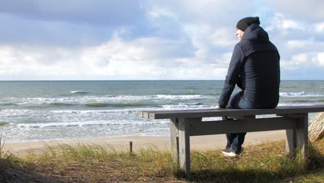 back view of caucasian male exploring nordic seaside forest, man sitting alone on the gray wooden bench on the beach, coastal pine forest, white sand beach, healthy activity concept, wide shot