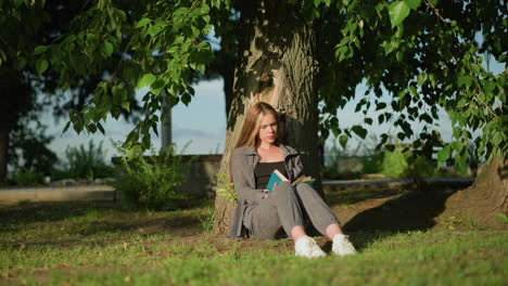 mujer sentada al aire libre con las piernas ligeramente estiradas, apoyada en un árbol en un campo cubierto de hierba, leyendo un libro bajo la cálida luz del sol, las hojas de los árboles se balancean suavemente en la brisa, el fondo presenta vegetación