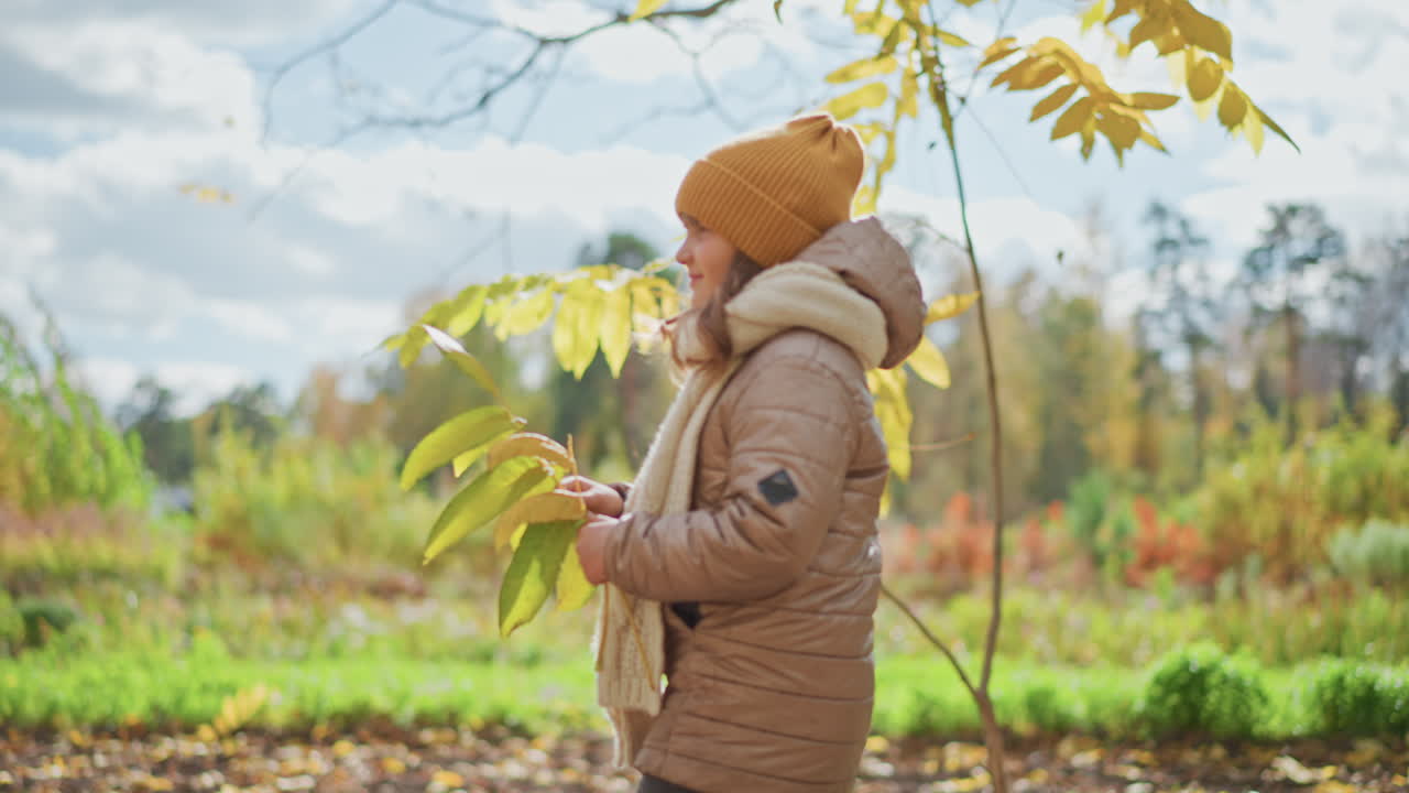 side view of kid walking through colourful autumn park path holding yellow leaf while wearing mustard beanie, beige jacket and knitted scarf under cloudy sky with vibrant foliage and gentle breeze