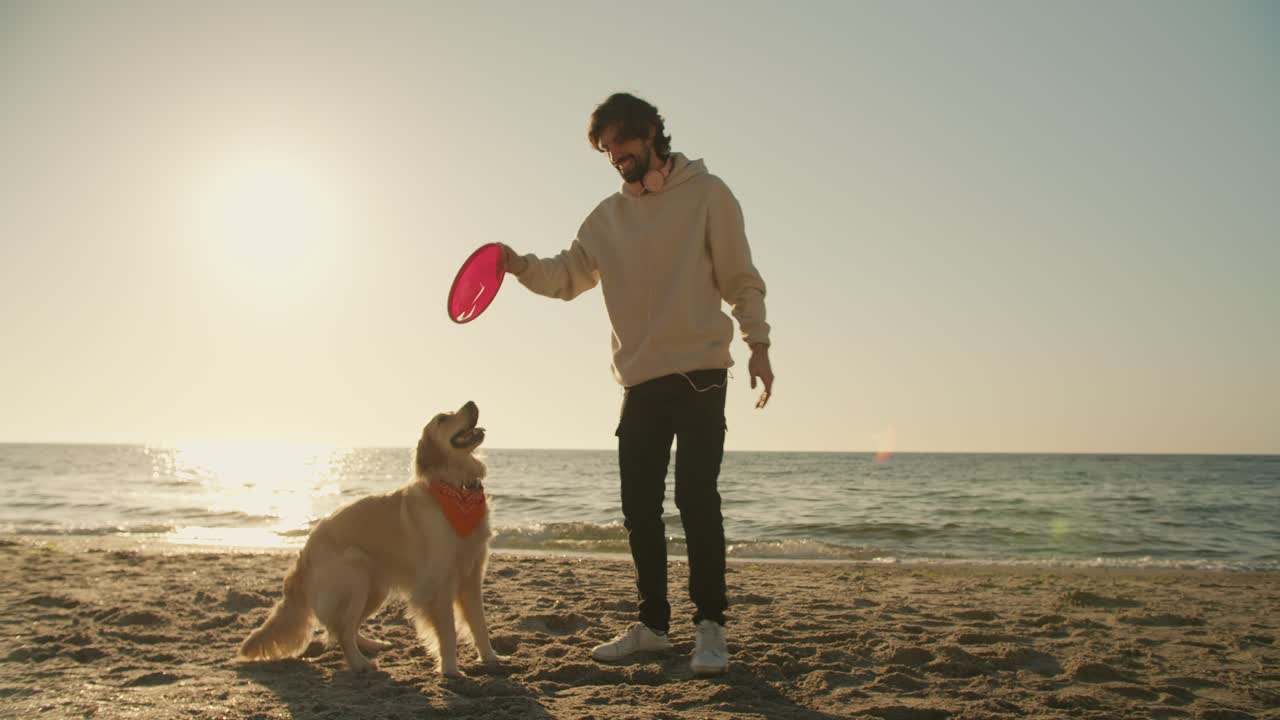 el tipo juega con un juguete rojo con su perro de color claro en una playa soleada por la mañana