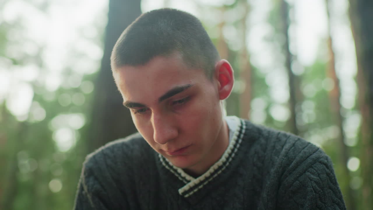 close up of young boy looking downward with serious expression while focusing on something outdoors in forest setting with natural light and blurred tree background