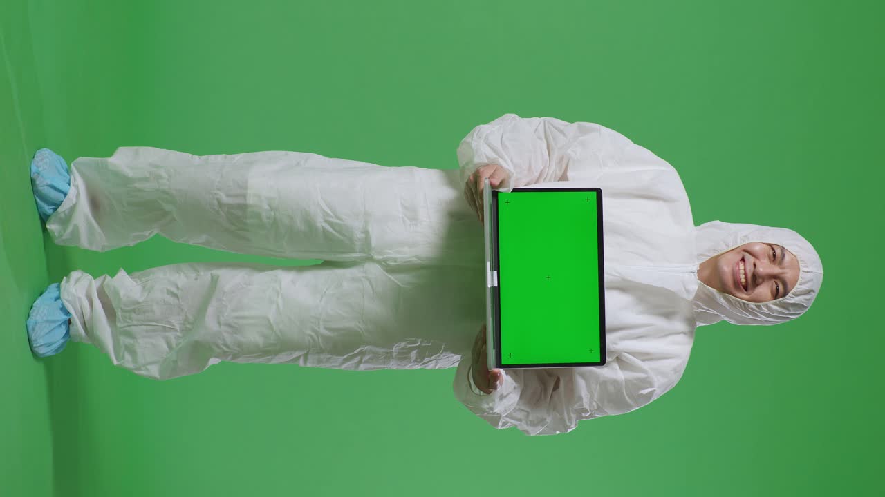 Scienziato maschio asiatico a figura intera che sorride e mostra un laptop con schermo verde mentre è in piedi su uno sfondo a schermo verde