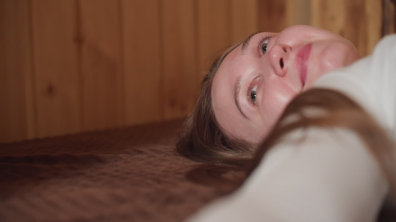 Close up of girl lying on bed, hand extended feeling soft texture of material, thoughtfully gazing upwards, serene and reflective moment in cozy indoor environment with warm wood backdrop