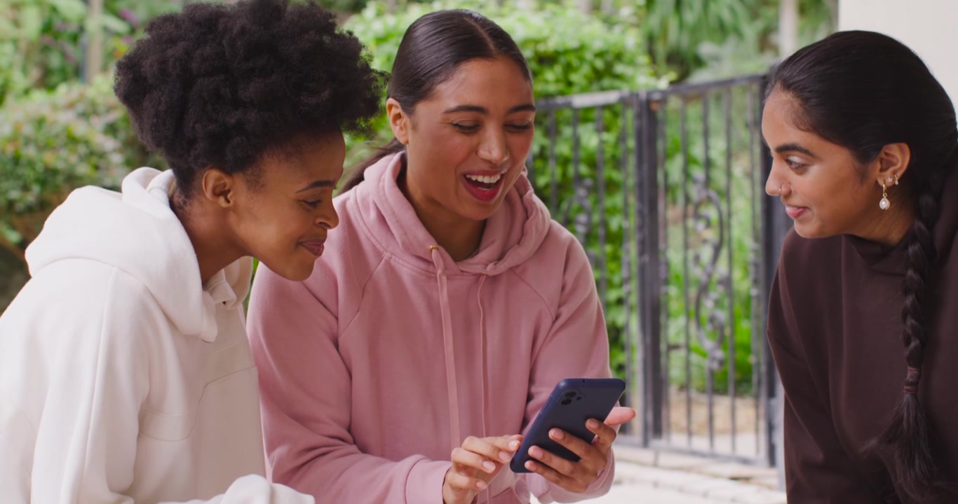 Diverse female friends sitting on porch, center woman tapping smartphone showing photos and talking