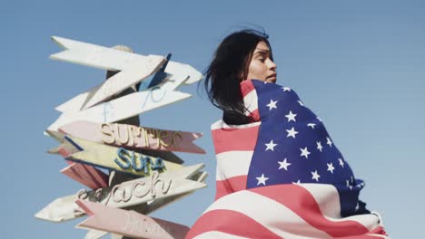 Feliz-Mujer-Hispana-Bailando-Con-Bandera-Americana-Junto-A-Un-Cartel-En-Una-Playa-Soleada,-Cámara-Lenta