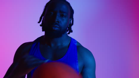 Studio-Portrait-Of-Male-Basketball-Player-Wearing-Team-Vest-Bouncing-And-Moving-Ball-Against-Colourful-Low-Key-Mixed-Lighting