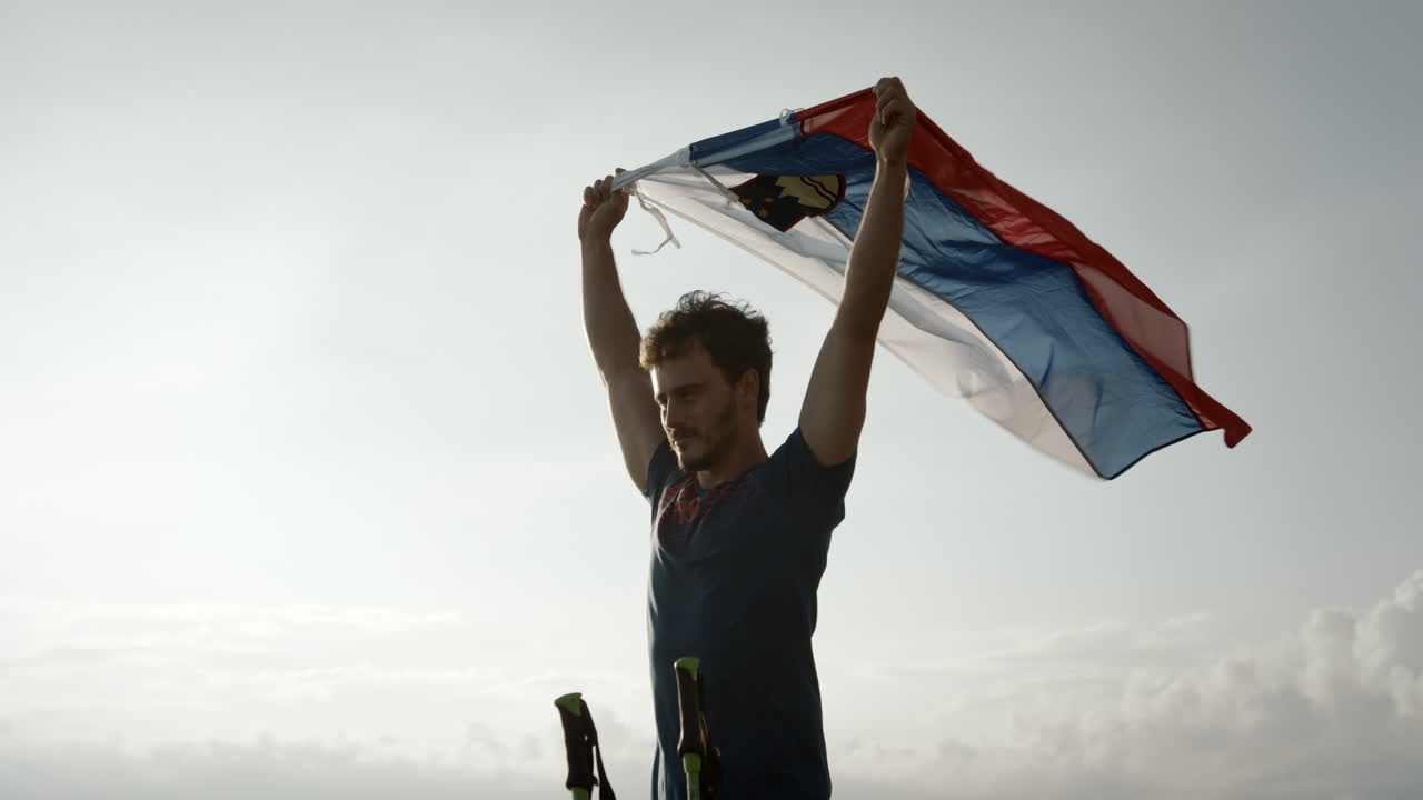 A young hiker holding in his hands a slovenian flag above his head and let it flutter in the wind, camera cicling arond him