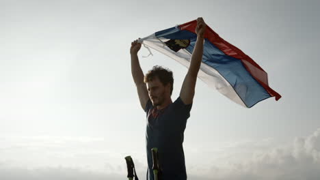 Un-Joven-Excursionista-Sostiene-En-Sus-Manos-Una-Bandera-Eslovena-Sobre-Su-Cabeza-Y-La-Deja-Ondear-En-El-Viento,-La-Cámara-Gira-A-Su-Alrededor