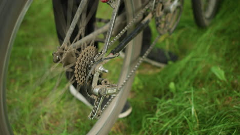 close-up of someone wearing black trousers and sneakers standing next to parked bicycle in grassy field, lifts the back tires and pedal, causing the tires to rotate, then gently lower the bike