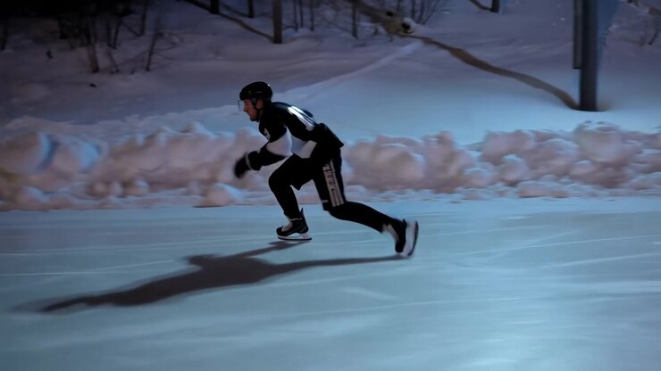 A solitary skater in dark winter clothing moves across an icy outdoor surface near a snowy embankment, with bare trees in the background under cold blue evening light