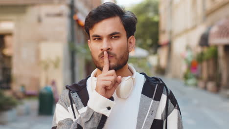 serious indian young man presses index finger to lips makes silence hush gesture in city street