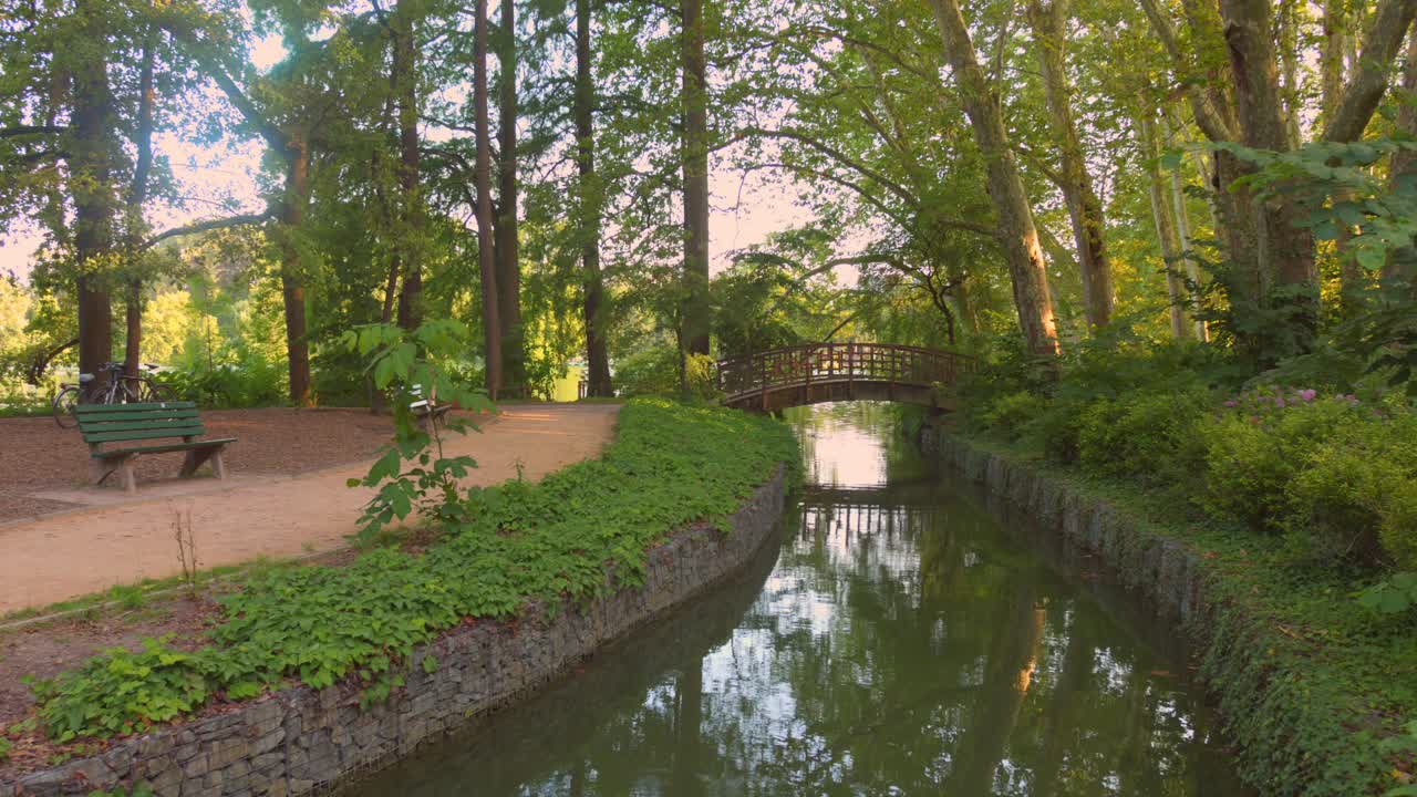 Wander through the tranquil Park of the Golden Head, lined with lush trees, as the soft river flows beneath a charming bridge and a welcoming bench awaits visitors nearby.