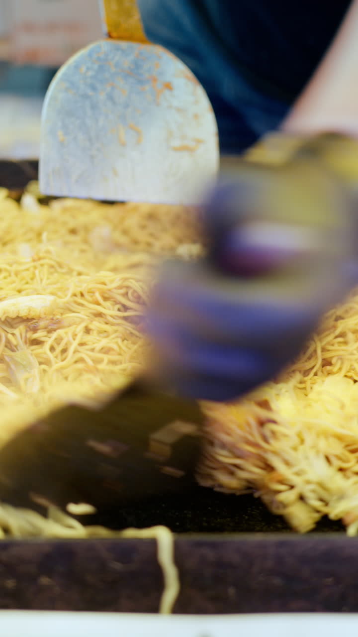 Close up of a vendor using metal spatulas to toss the fried noodles around on the teppan at a street food market in Japan. Vertical
