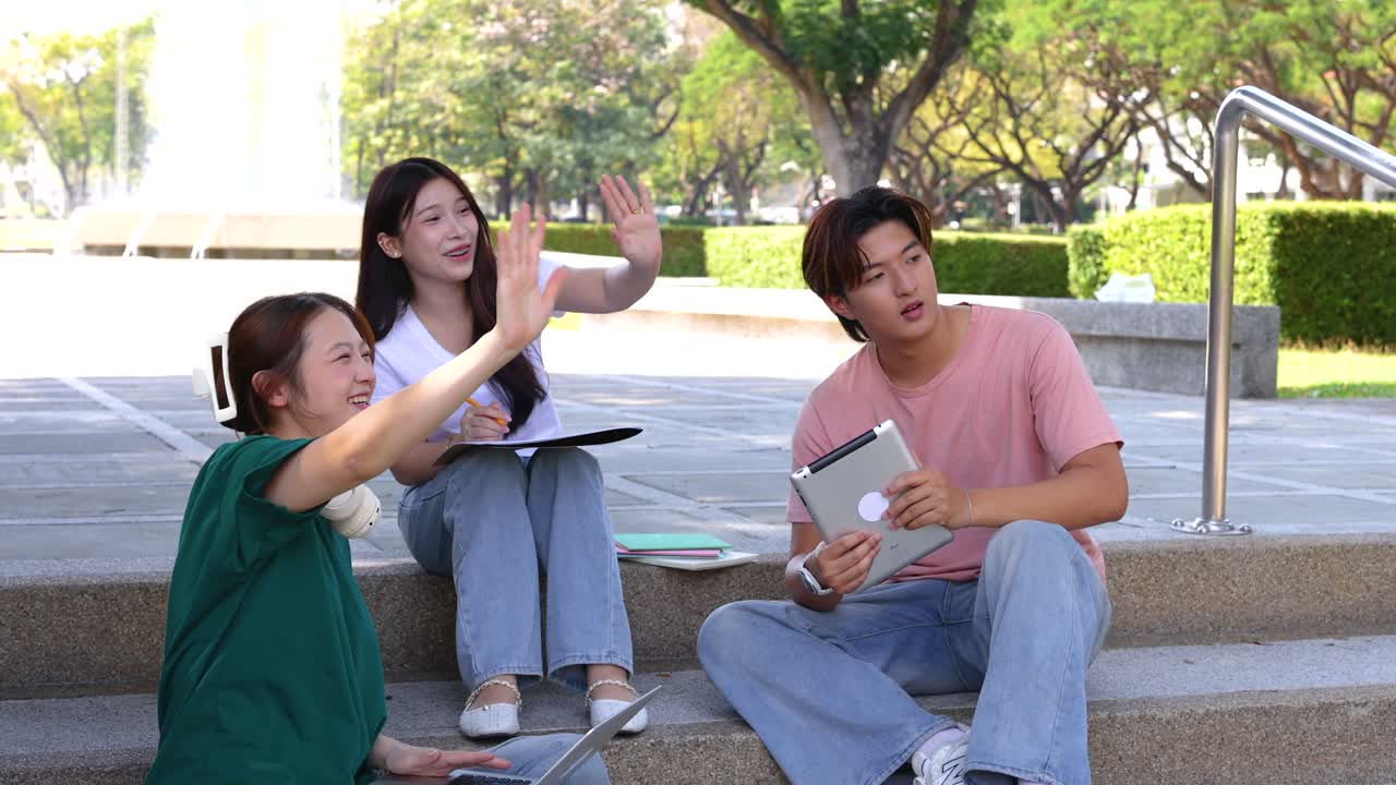 3人のタイ人学生が、キャンパスの階段に座ってタブレットやラップトップを使いながら、友人に手を振っています。
