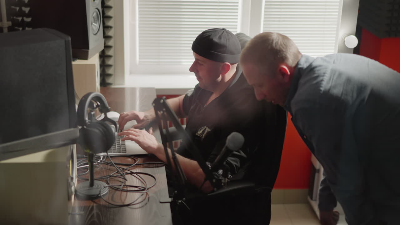 두 남자가 스튜디오 방에 앉아 있는데, 한 남자는 컴퓨터 앞에 앉았고, 아마도 프로듀서였고, 검은 옷을 입고 있었고, 다른 한 남자는 긴 팔을 입은 셔츠를 입고, 그의 무을 손으로 고 있었다.