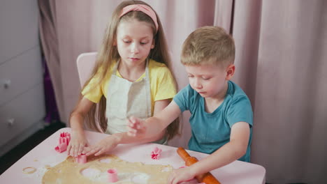 girl in yellow shirt concentrating on cutting dough using pink cutter while boy beside her proudly holds and shows his cutout shape, creating lively baking moment with rolling pin and flour on table