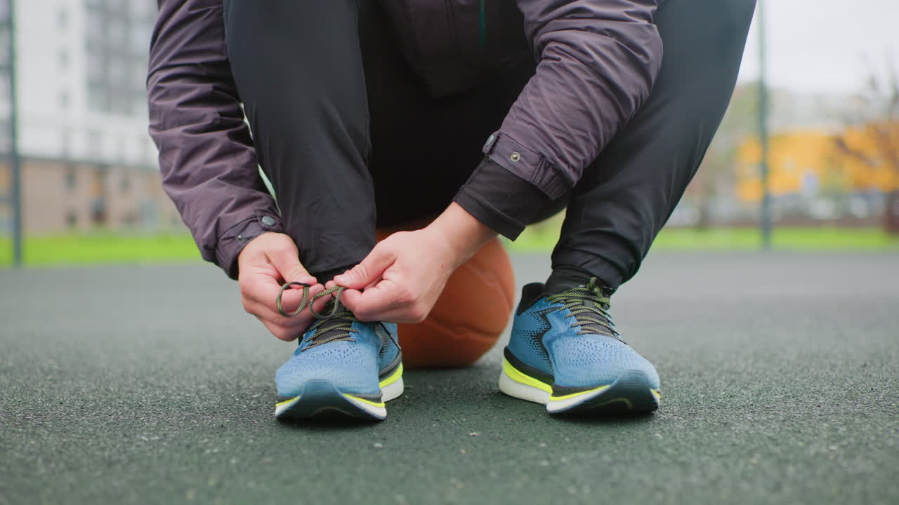 Corridore che si allaccia le scarpe da ginnastica vicino al campo da basket, primo piano delle mani che allacciano i lacci inginocchiandosi sull'asfalto con il pallone da basket sotto il ginocchio, fantastica luce mattutina urbana, rituale prepartita concentrato, campo grintoso
