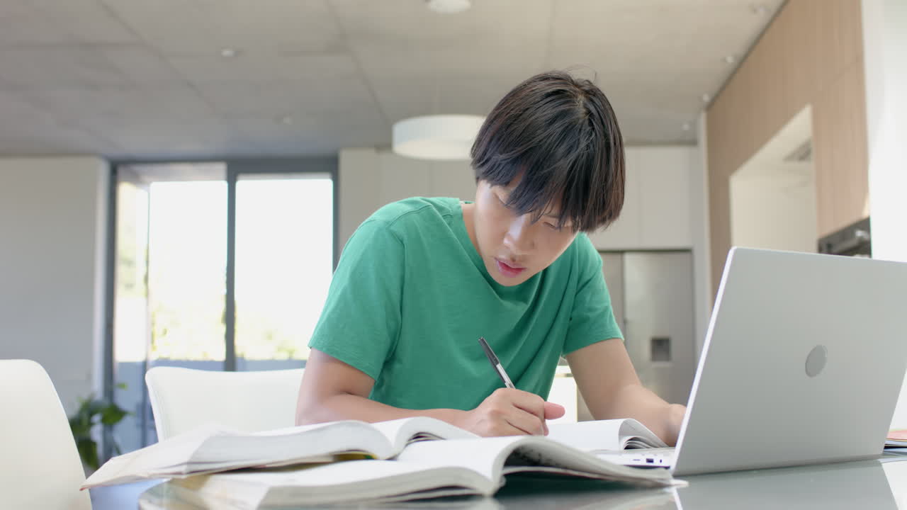 テーブルでノートパソコンを使って熱心に勉強するアジア系の10代の少年。