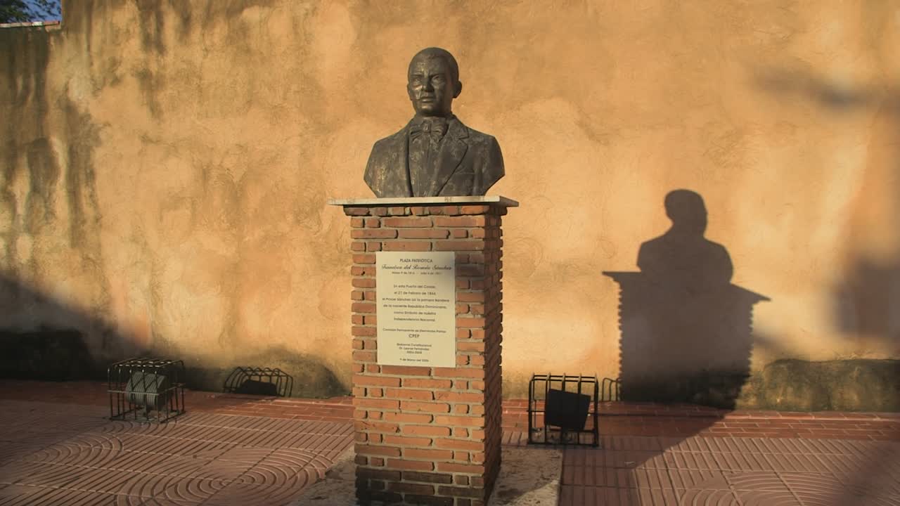 Inquadratura media di un monumento a busto su piedistallo di mattoni nella piazza patriottica di Santo Domingo, fotografato contro un muro ocra illuminato dal sole con forte ombra.