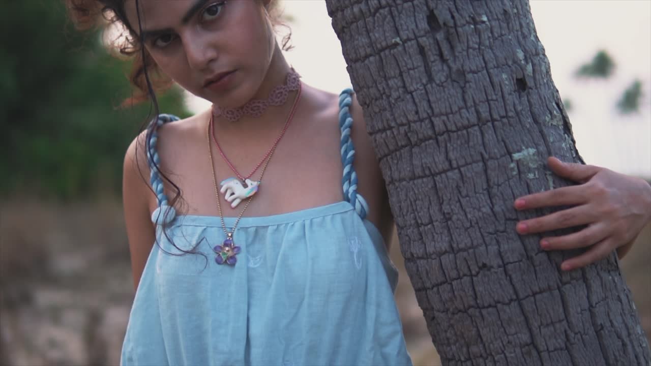 해변에서 줄기를 껴안고 카메라를 위해 포즈를 취하는 현대적인 파란색 드레스를 입은 아름다운 소녀