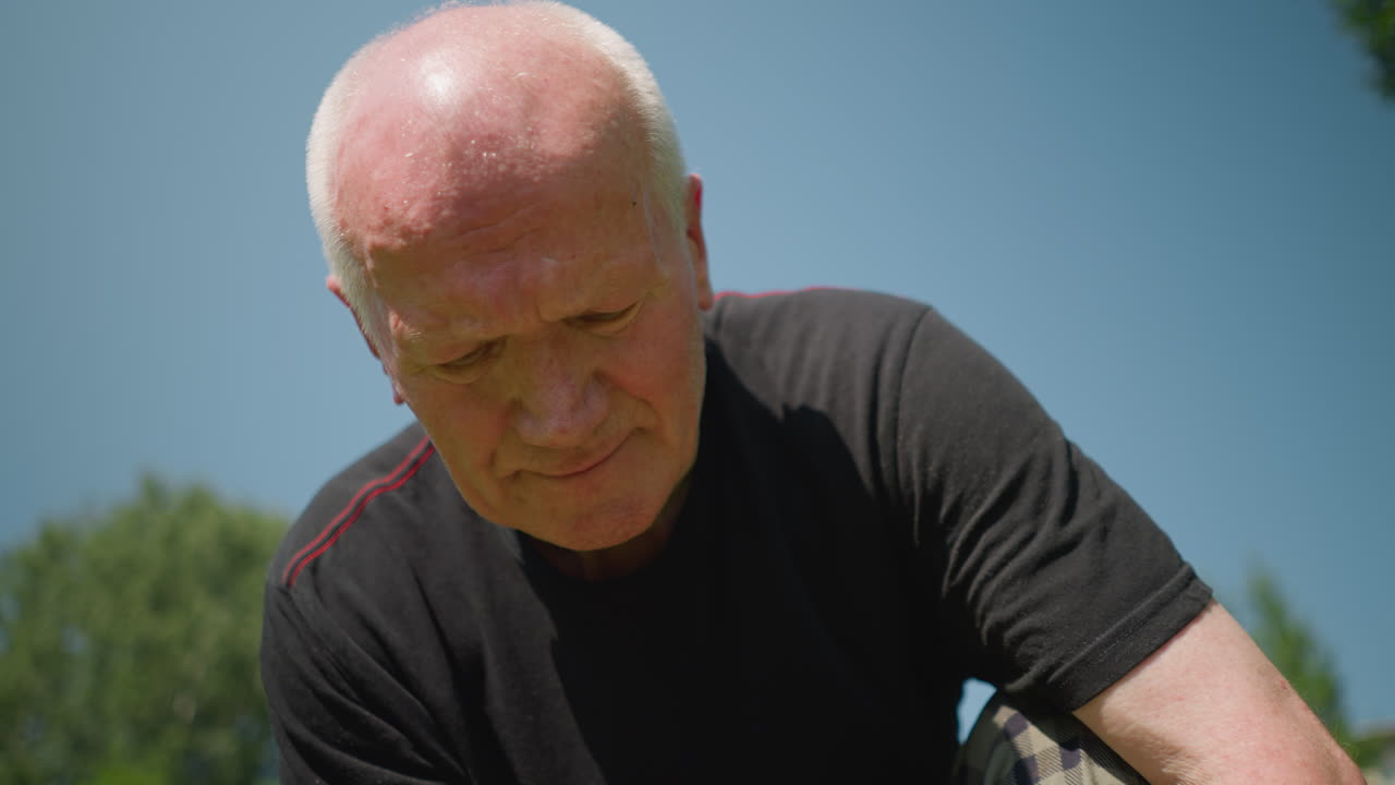 An elderly man is kneeling down, deeply focused on working on something as he engages in his task with a blurred view of trees in the background