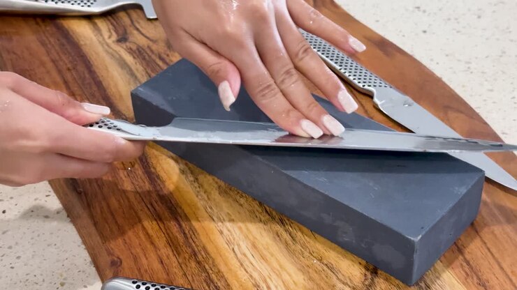 A person sharpens a chef knife on a whetstone atop a wooden cutting board, using smooth, controlled strokes under bright, even kitchen lighting