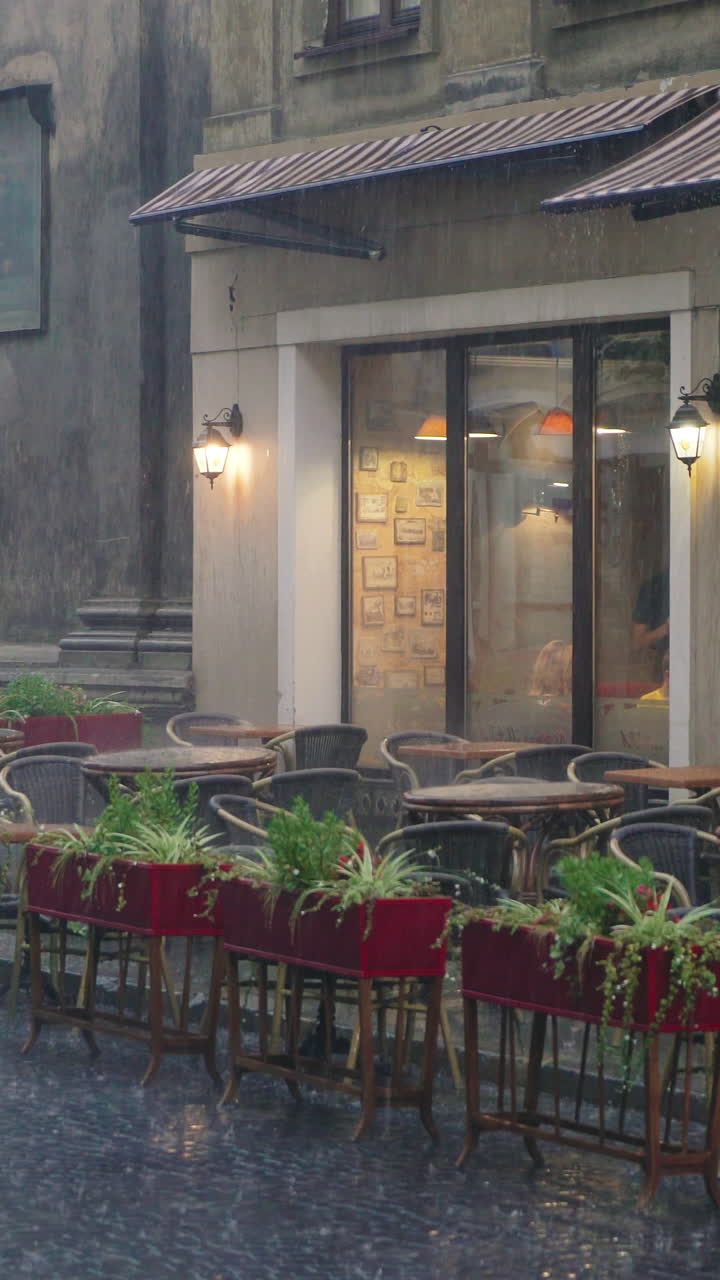 raindrops are falling on the tables and chairs of the restaurant on the street. Wet summer terrace with flower pots is near the road. Vertical video