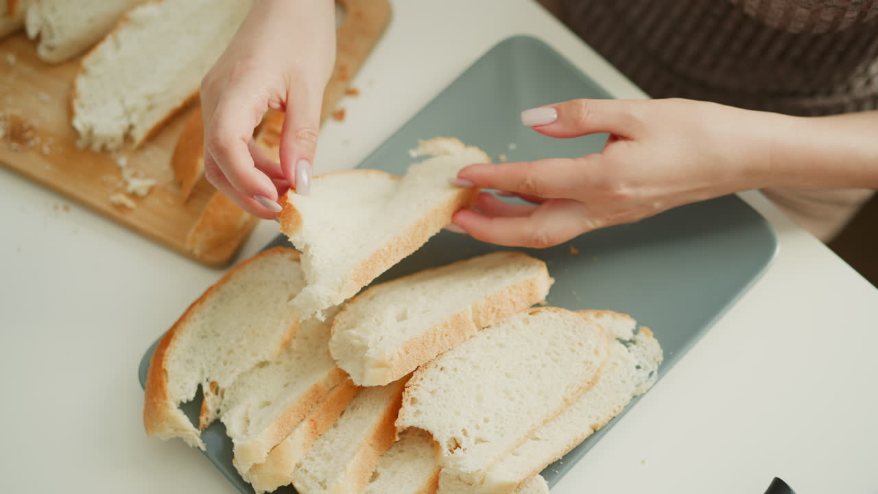 Close up of bread slices being arranged on tray by female hand on white table in bright kitchen morning light, highlighting careful process, texture of crust, crumbs on wooden board and knife nearby