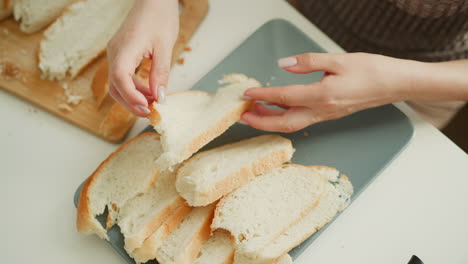 close up of bread slices being arranged on tray by female hand on white table in bright kitchen morning light, highlighting careful process, texture of crust, crumbs on wooden board and knife nearby
