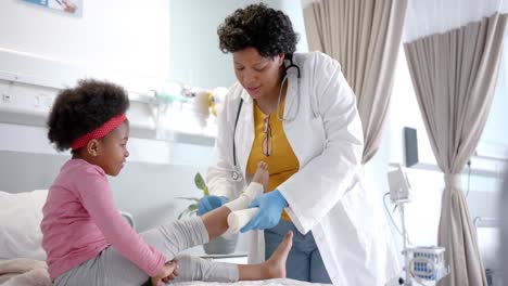 African-american-female-doctor-putting-bandage-on-girl-leg-in-hospital-room,-slow-motion