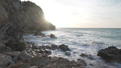 Panorámica-De-La-Cámara-A-La-Derecha,-Costa-Rocosa-Del-Mar-Temprano-En-La-Mañana-Cuando-Las-Olas-Llegan-A-La-Costa-Y-La-Llamarada-Solar-Golpea-La-Lente